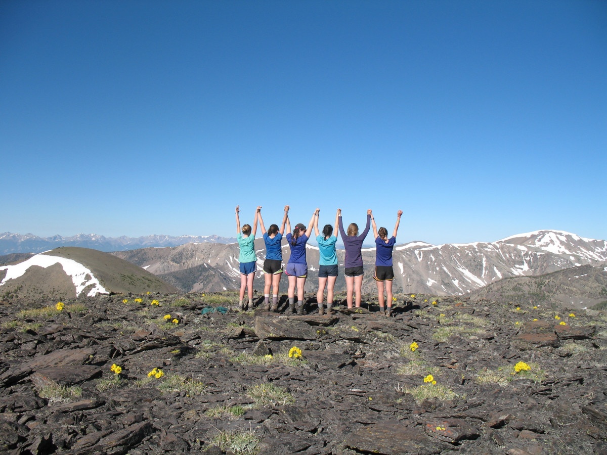 Six teenage girls stand together on a rocky expanse in the mountains and raise their arms up holding hands