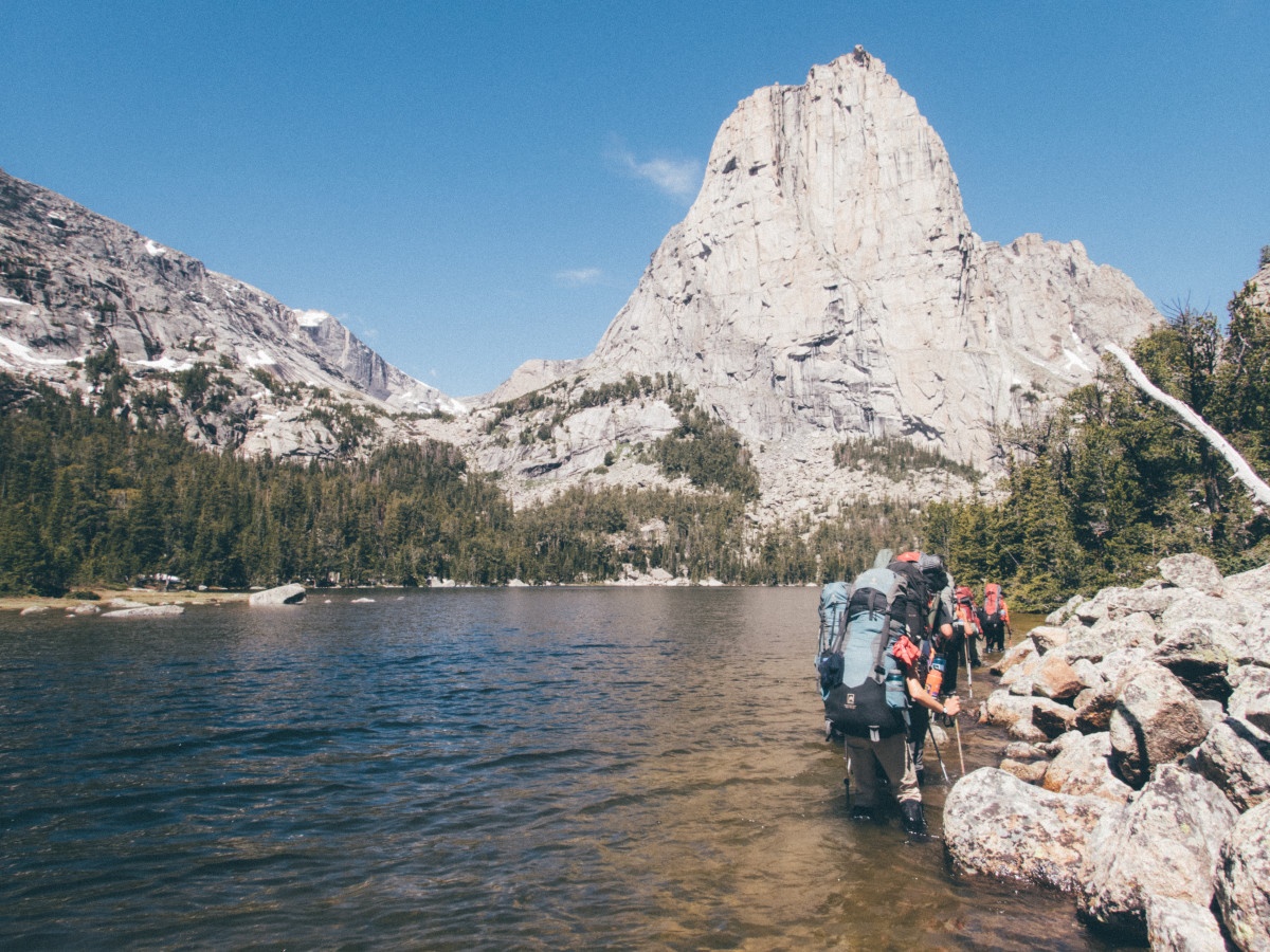 students wearing backpacks wade single file along the edge of a lake, using trekking poles for balance