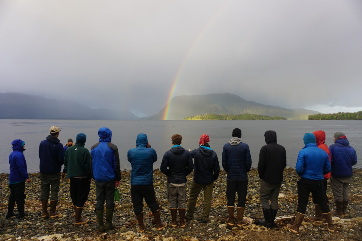 group of NOLS participants stand on a rocky beach looking out at a rainbow coming out of the mist