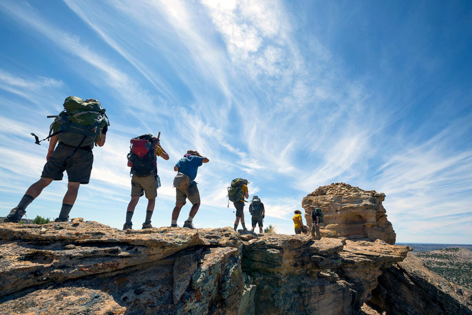 line of hikers wearing backpacks walk single file along a rocky outcrop