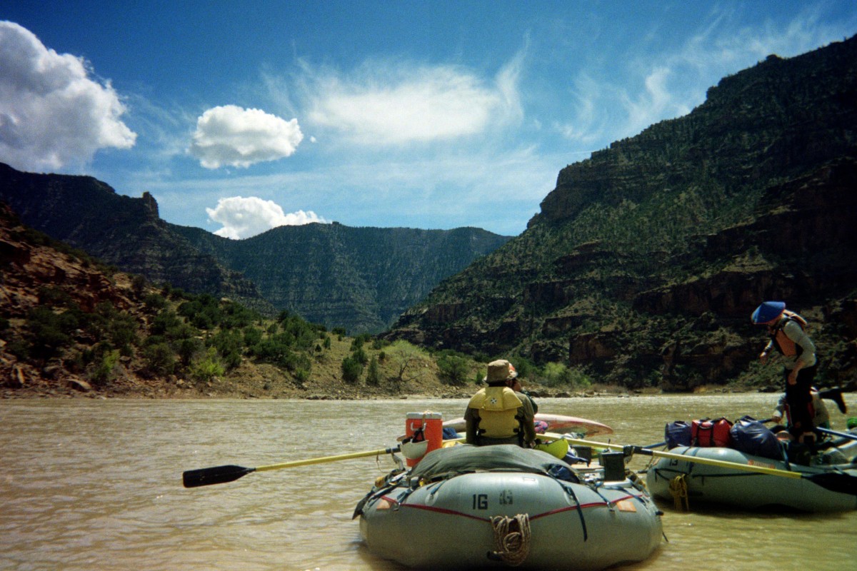 Students in two rafts with oars on a muddy river with tall canyon walls