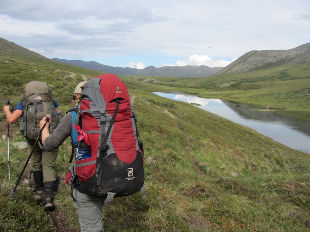 two students wearing backpacks use trekking poles to hike across a grassy slope in Alaska with glassy river to the right