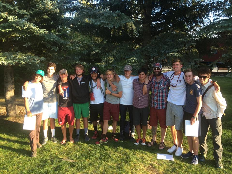 12 male NOLS students stand in a line with arms draped around each other on green grass after their course