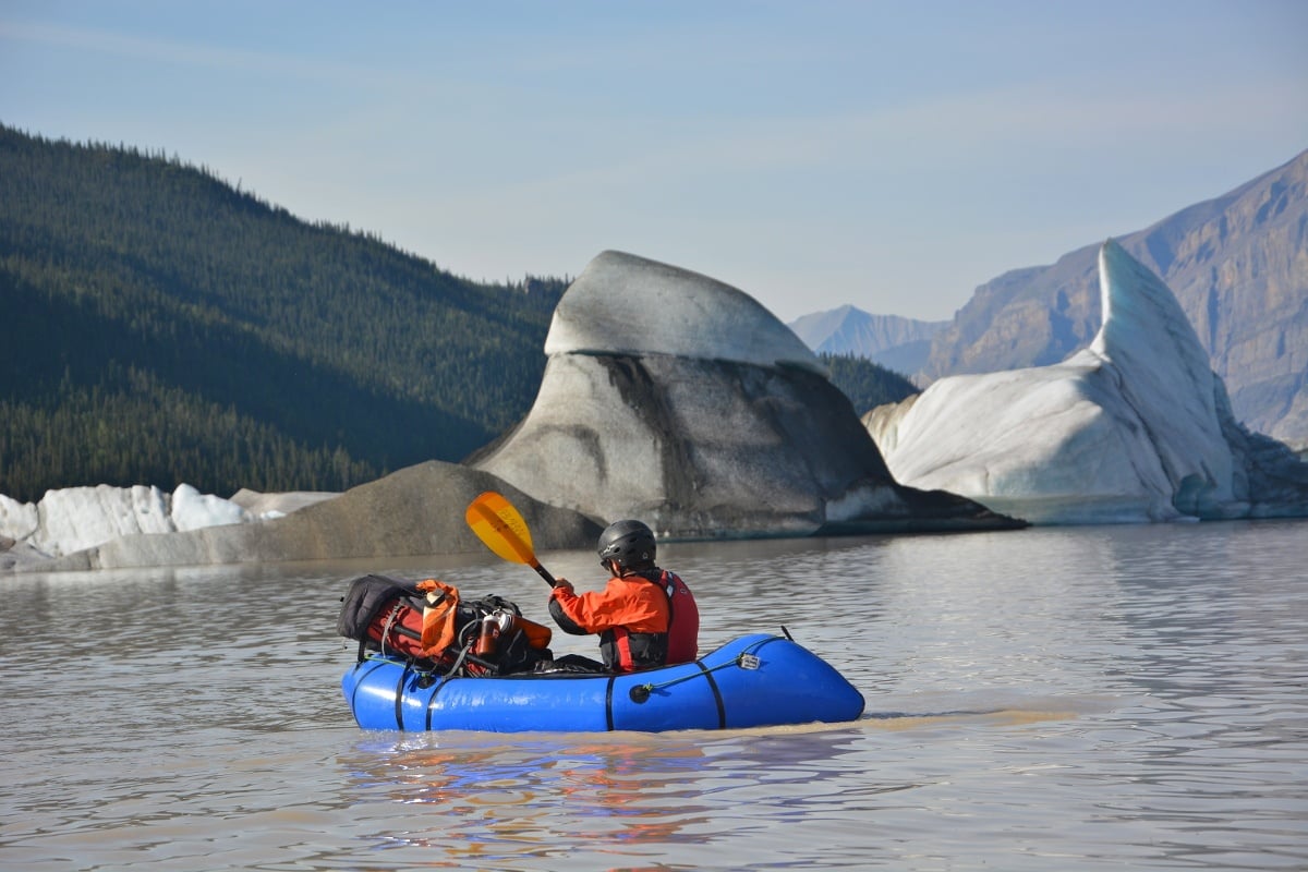 NOLS student paddles blue packraft on calm water past dirt encrusted icebergs