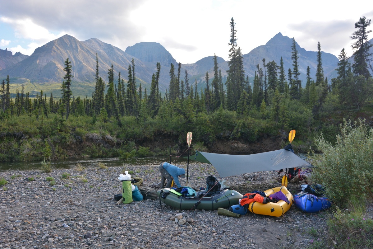 packrafting camp with tarp strung up between kayak paddles on rocky ground near a river in Alaska's mountains