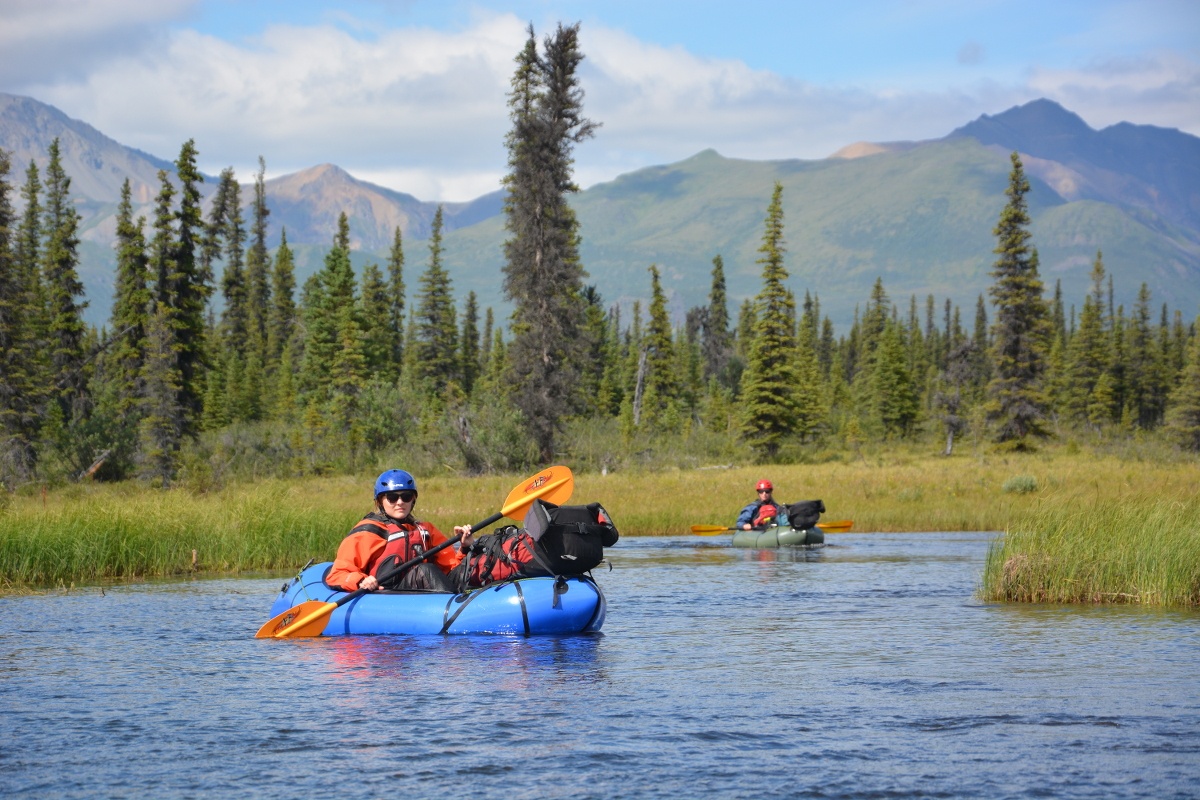 smiling student paddles a blue packraft on flat water beside pines in Alaska's mountains