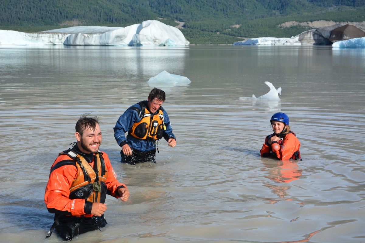 three NOLS students wearing drysuits stand in murky water with floating ice