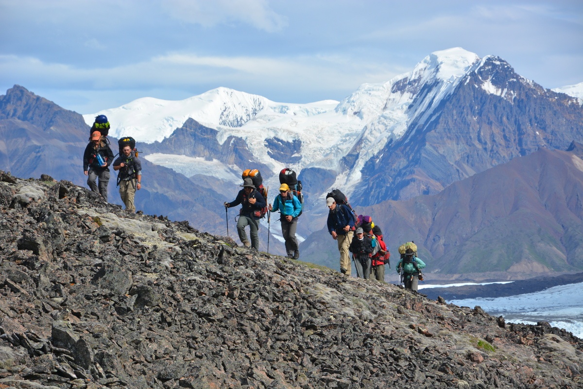 backpackers, some carrying trekking poles, hike up a rocky slope with snowy mountains behind them