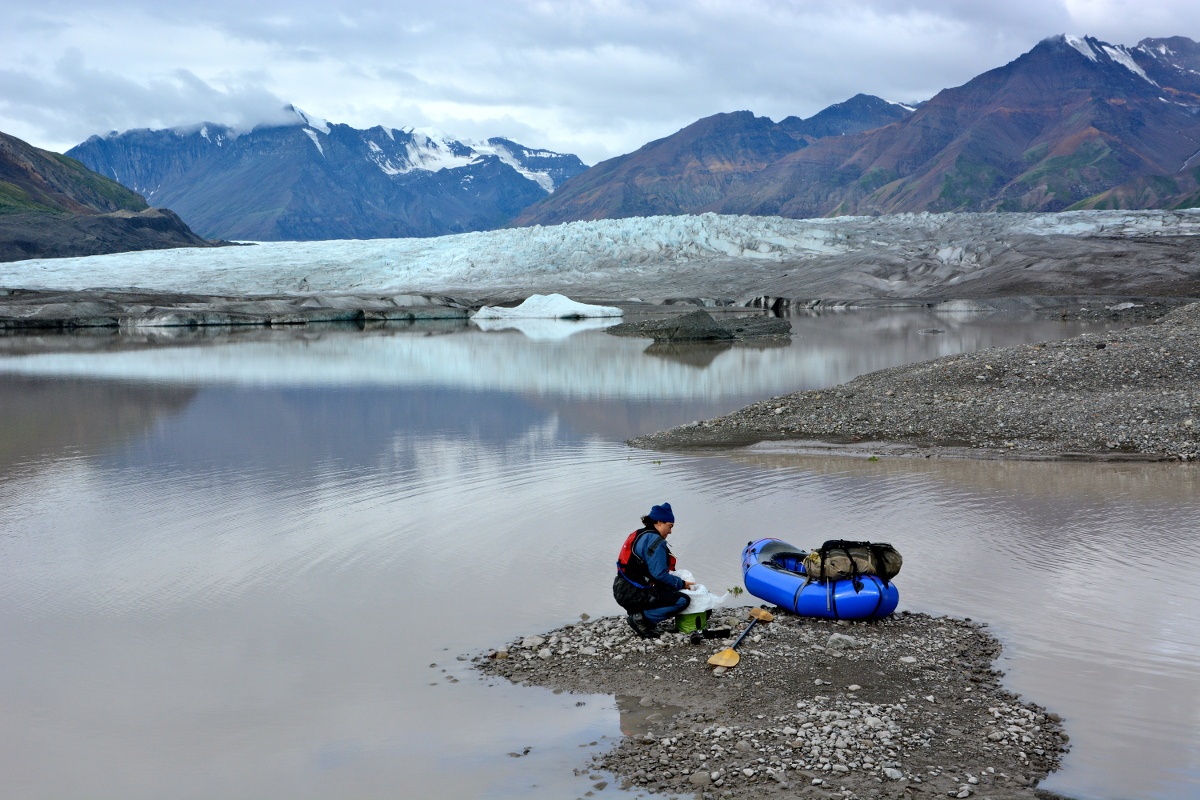 NOLS instructor packs up blue packraft on a rocky spit of sand near a glacial lake in Alaska