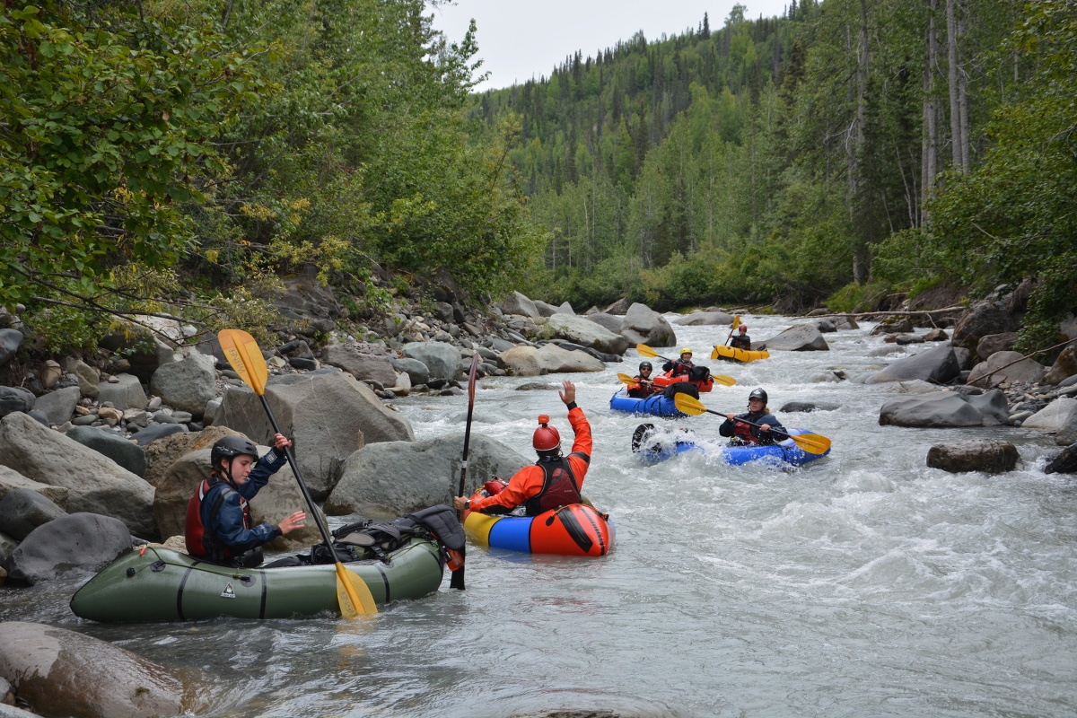 NOLS students wearing life preservers and helmets paddle packrafts down a small rapid on a river bordered by thick trees