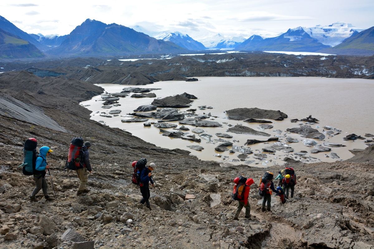 Hikers descending a talus field toward a muddy lake in Alaska's mountains