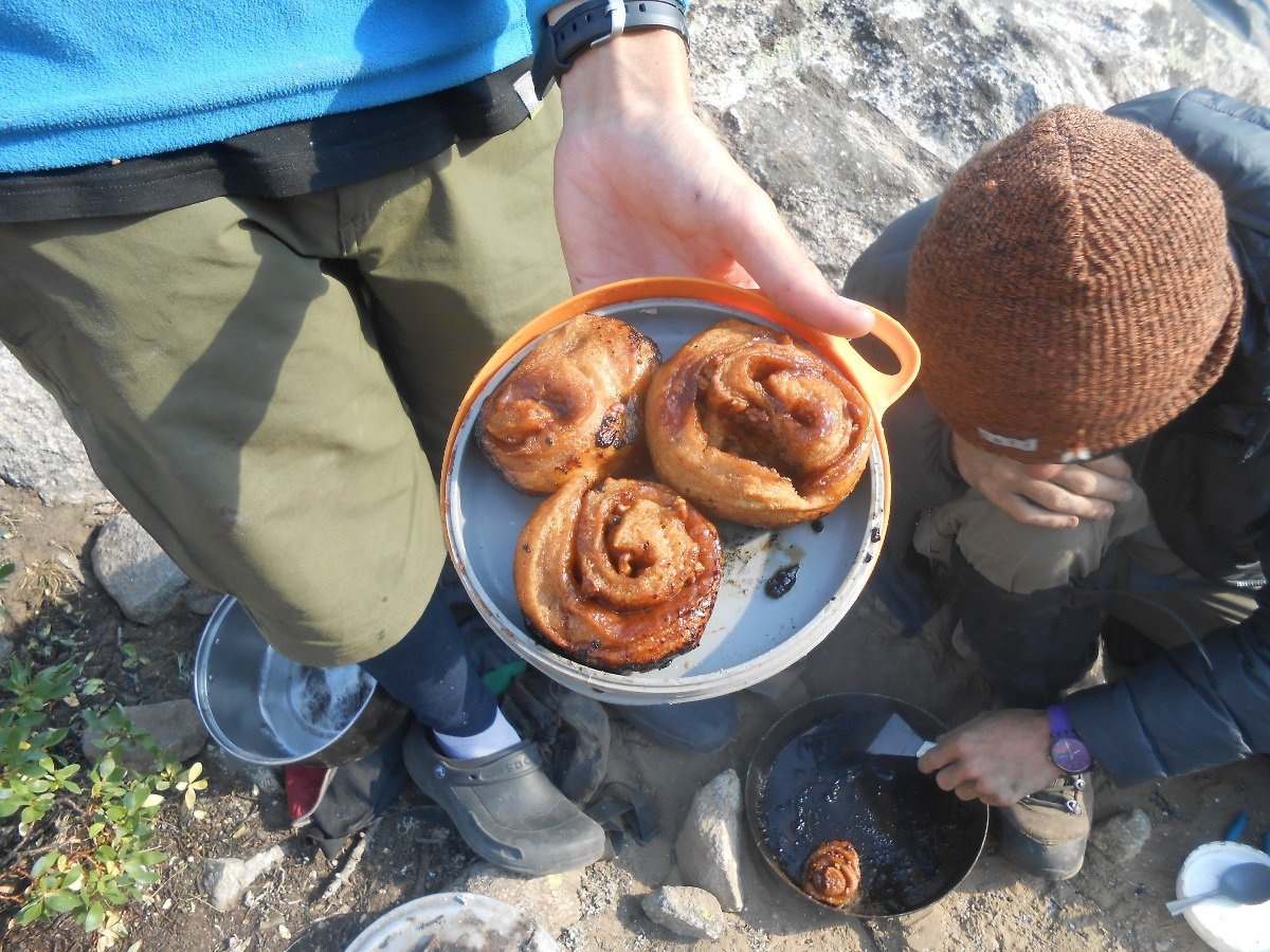 student holds pan of three cinnamon rolls, while another student squats on the ground, scraping a pan containing another roll