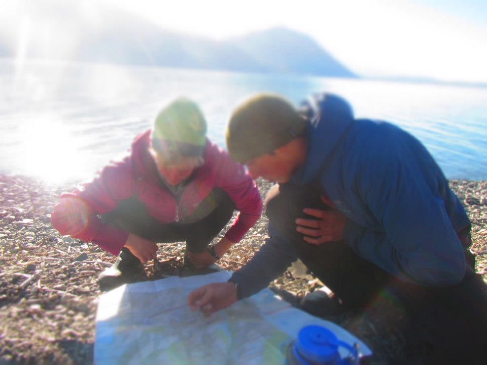 Two students crouch as they read a map on the shore of a body of water