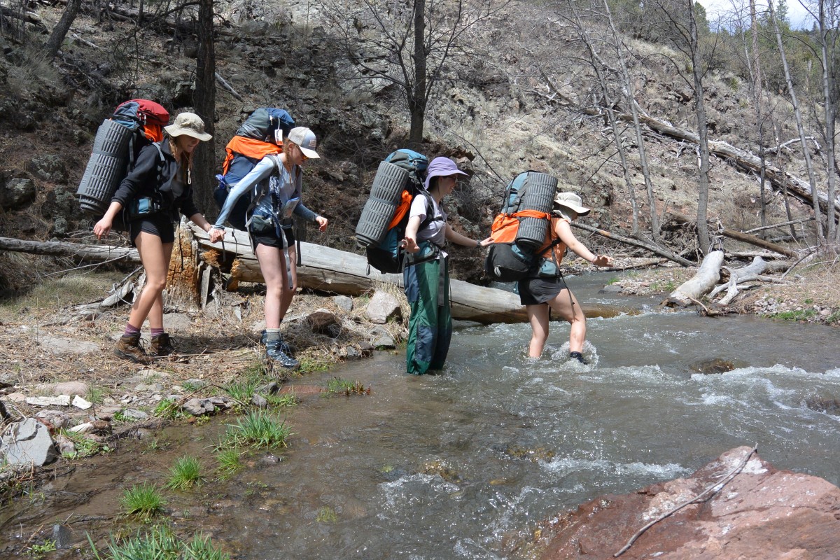 Four girls wearing backpacks help each other cross a creek