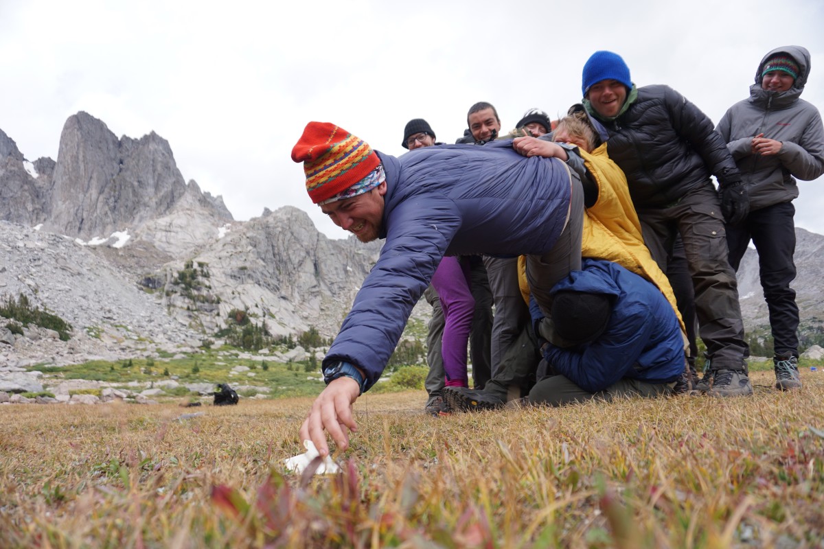 Group of people holding onto a person reaching to grab an object off the ground while team building in the mountains