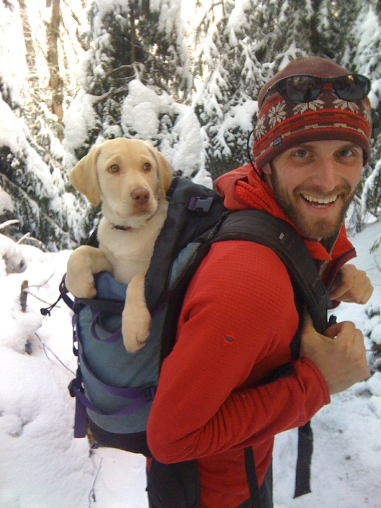 Smiling man wearing red fleece and hat carries yellow lab puppy in a backpack