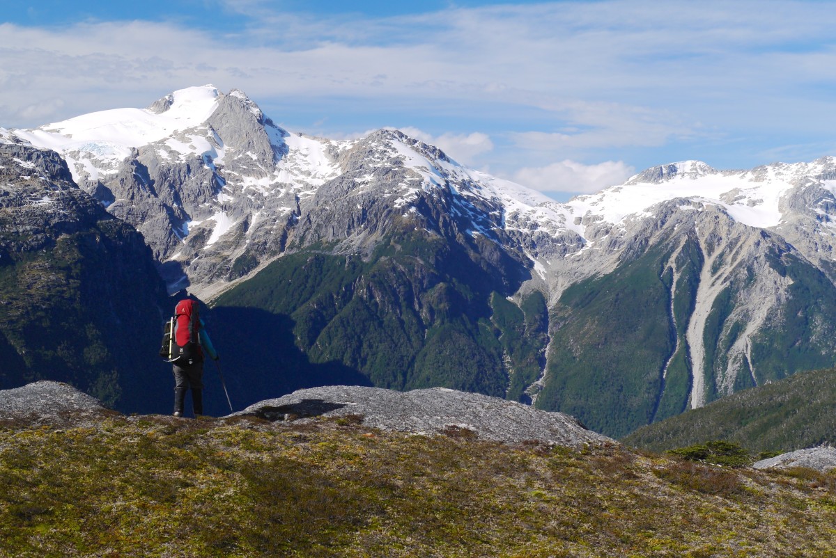 A backpacker pauses on top of the ridge with snowy peaks in the distance