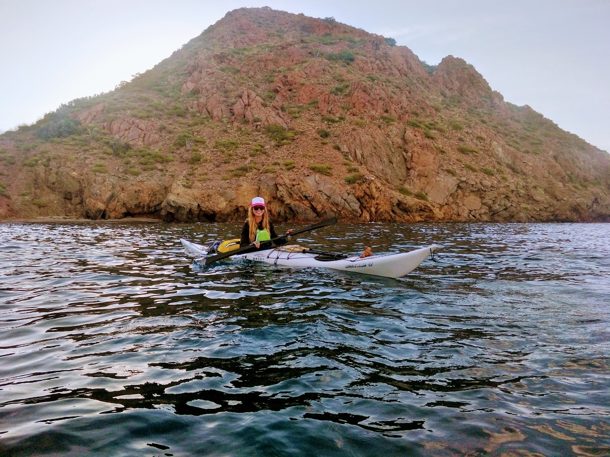 woman wearing baseball cap paddles white kayak near shoreline with steep rocky hill