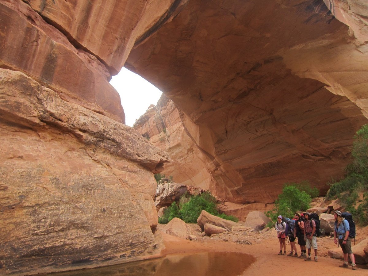 NOLS students stand by a shallow pool near a massive red rock arch in a canyon