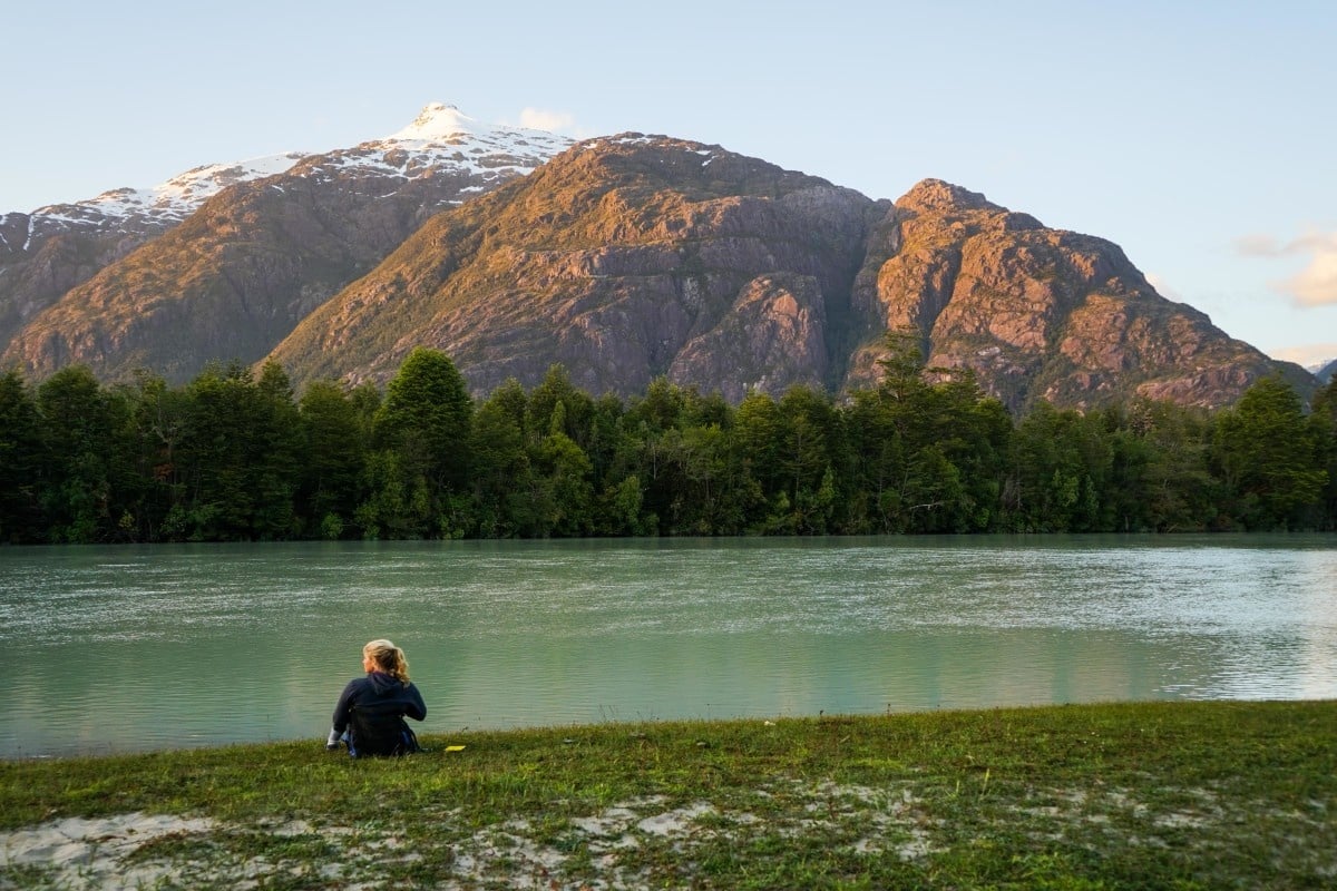female NOLS student sits in the grass looking across a river toward thick forest and mountains