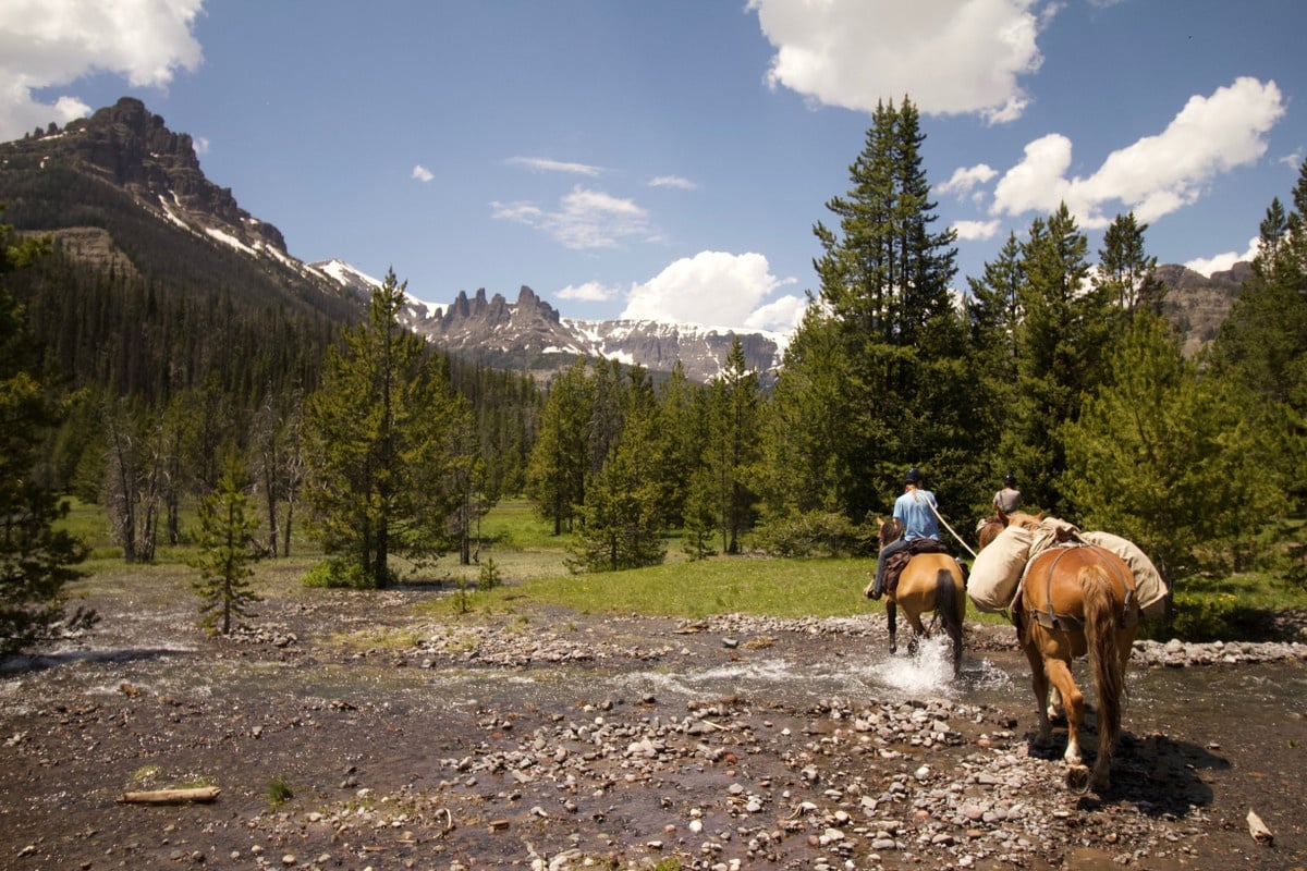 A rider and packhorse cross a stream with mountain ridges in the background