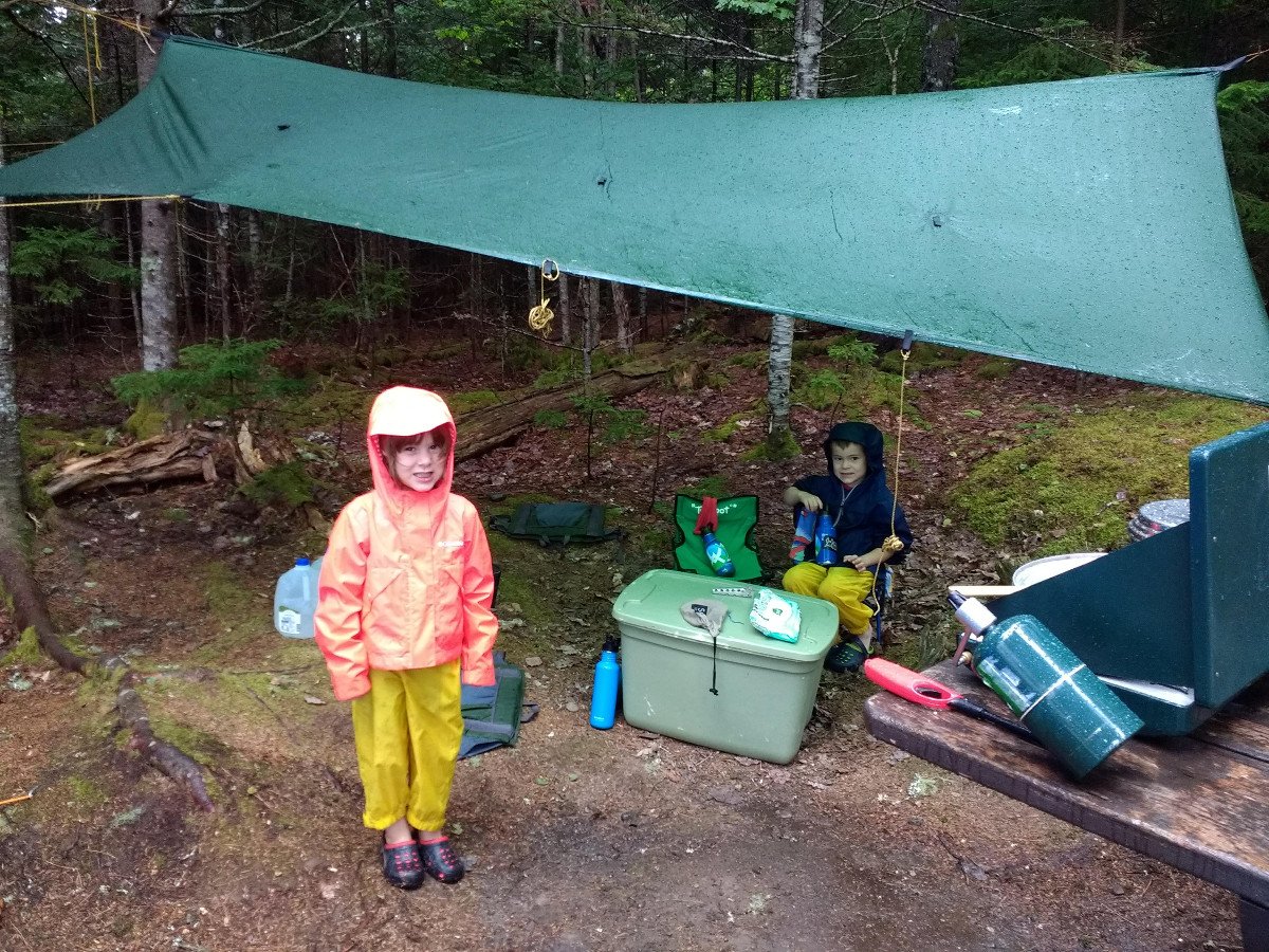 2 kids wearing raincoats under a tarp in camp