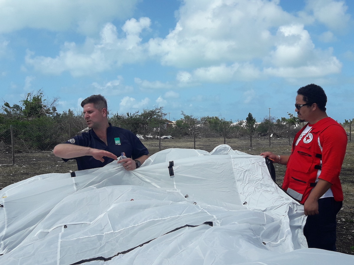 man wearing blue shirt helps man in Red Cross shirt set up white tent