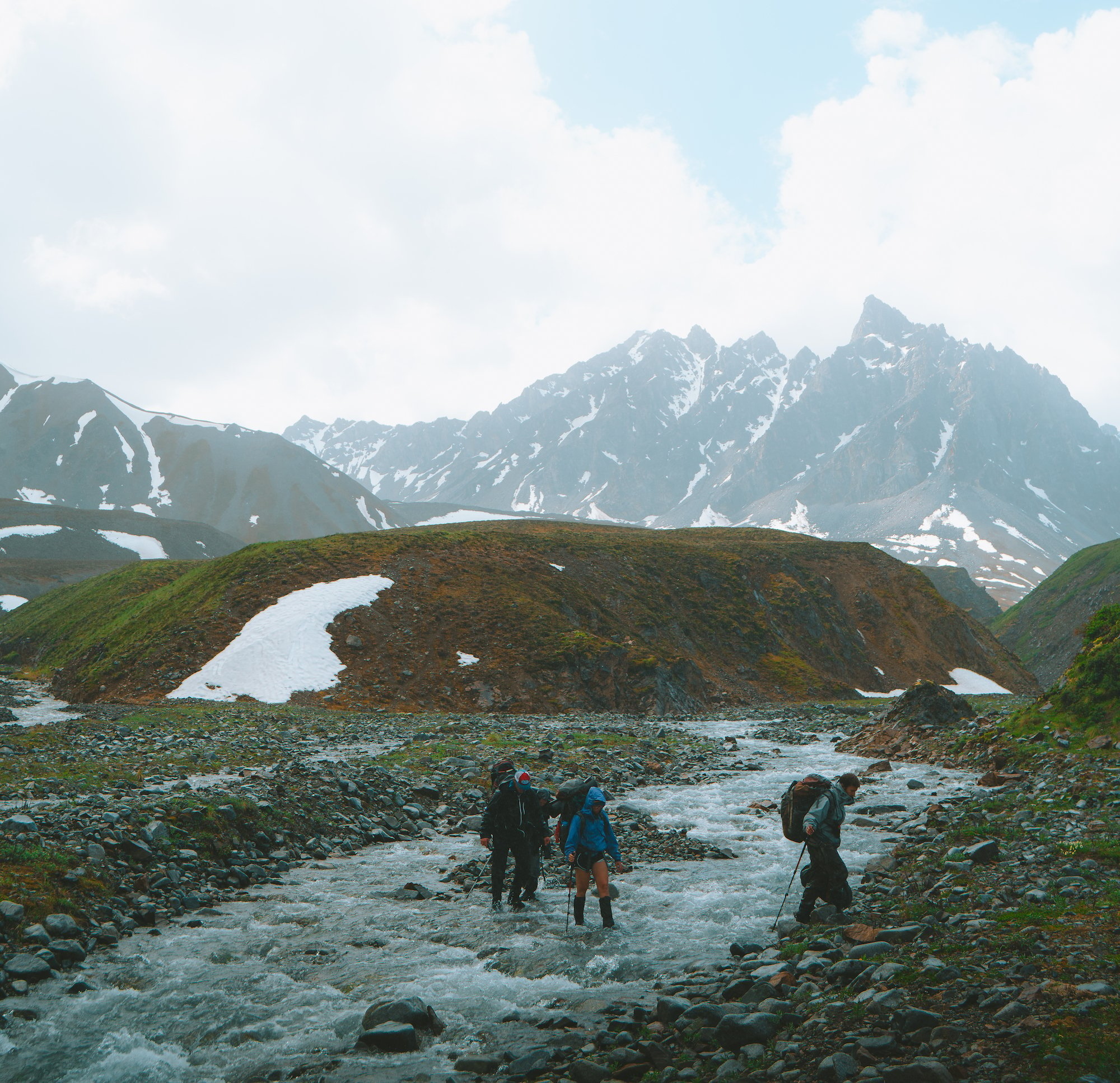 NOLS students cross a stream in mountains with patchy snow