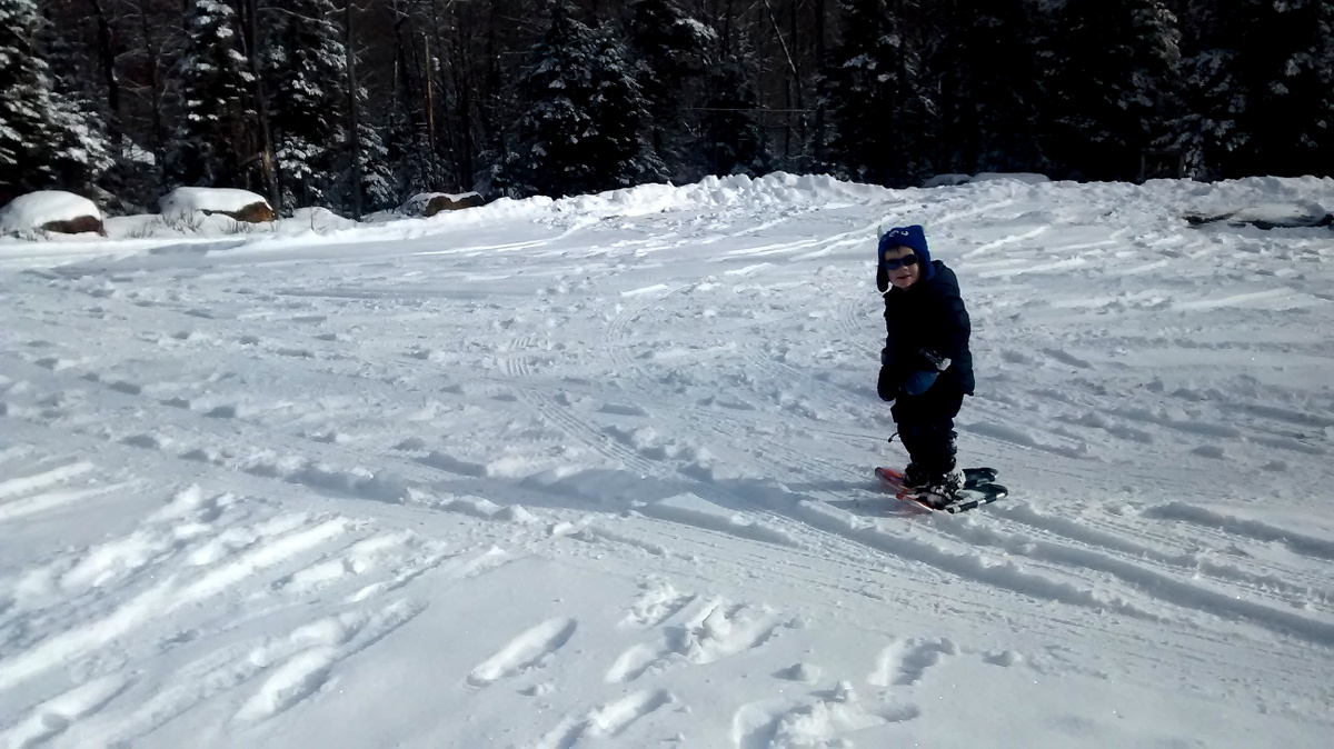 Smiling child wearing winter gear, sunglasses, and snowshoes stands in a snowy parking lot facing the camera