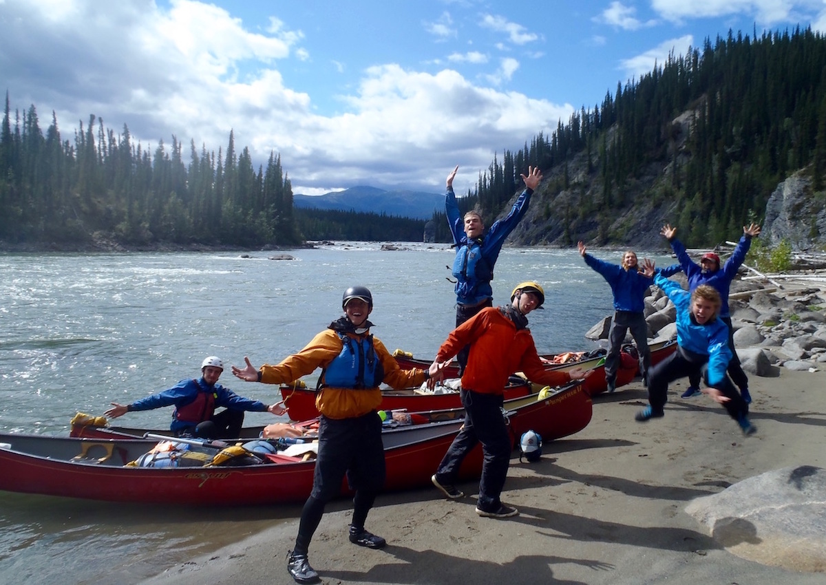 group of NOLS students make goofy faces and pose beside canoes and river in the Yukon