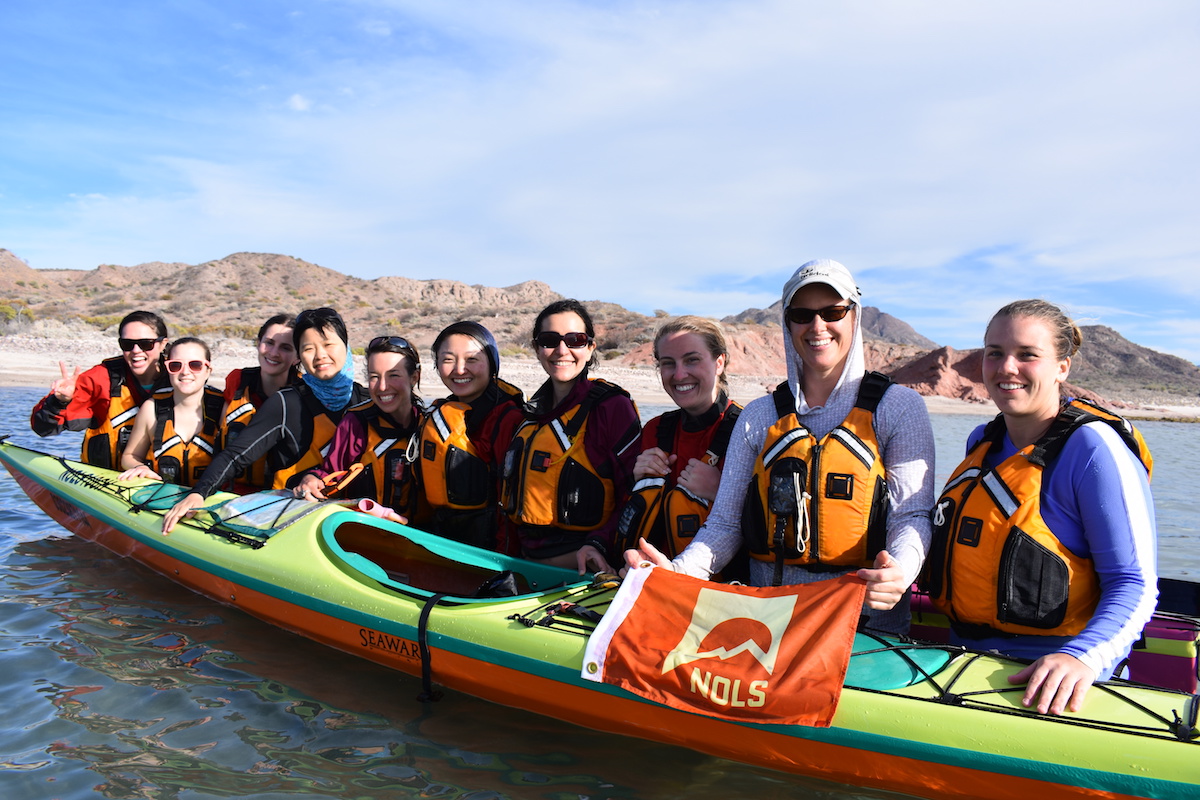 smiling female students stand in the ocean near sea kayak holding an orange NOLS flag