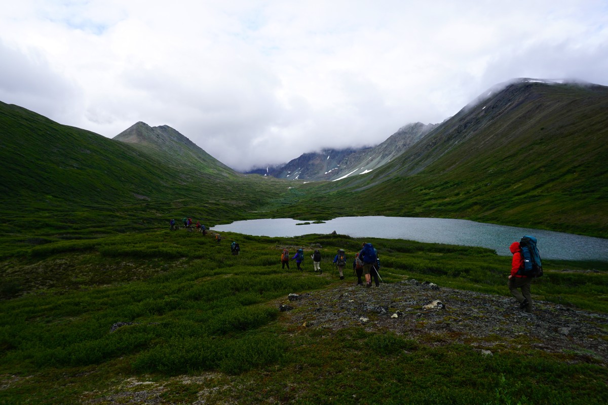 Backpackers hike along a lake in a lush mountain valley