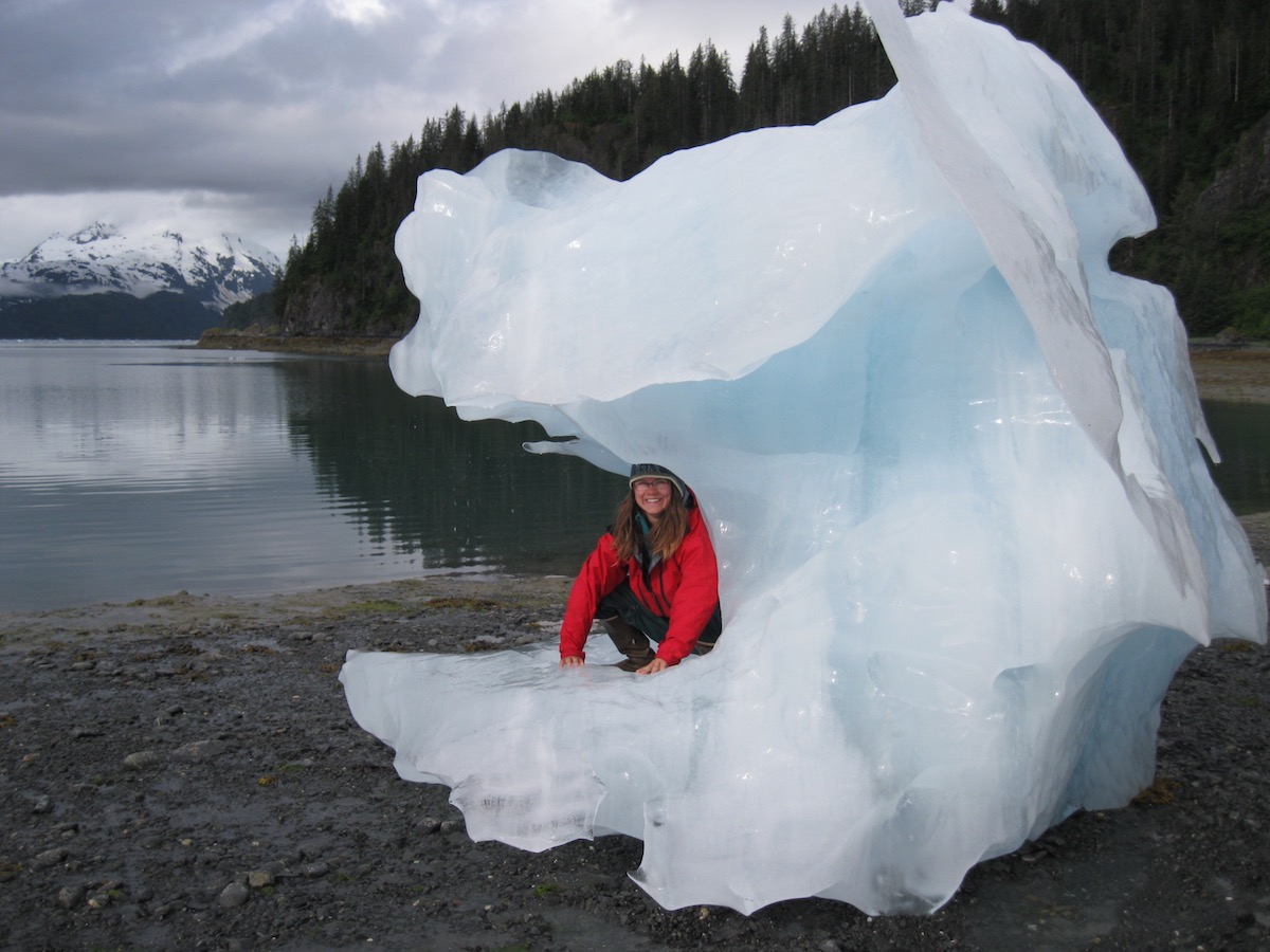 smiling person in red jacket kneels inside a large ice formation on the shore in Alaska