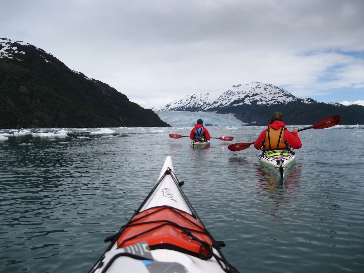 two paddlers sea kayak in Alaska on calm water with floating ice as seen over the nose of a kayak