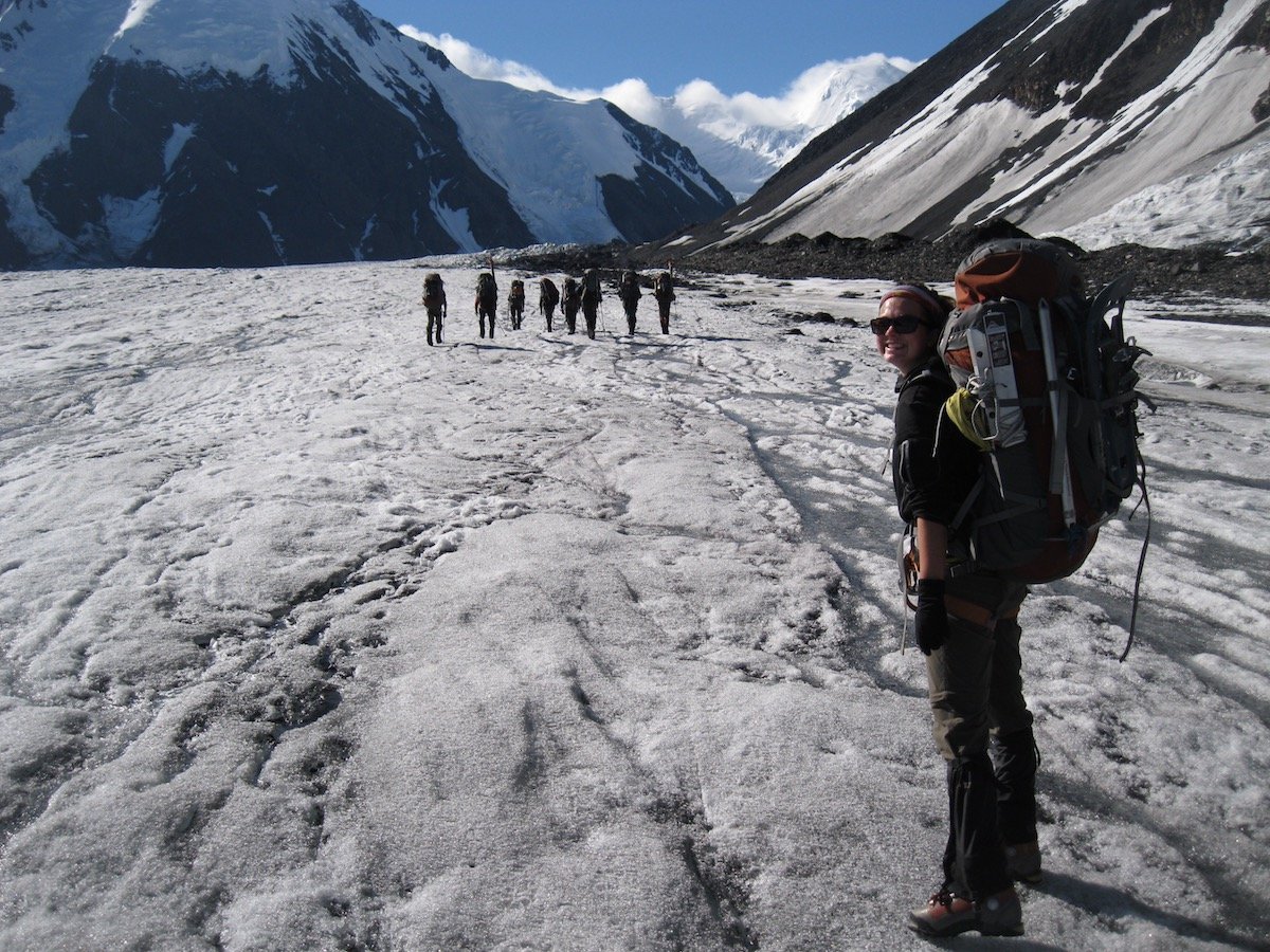 smiling woman wearing a backpack walks up a snowfield in Alaska with a group of eight people hiking in the distance