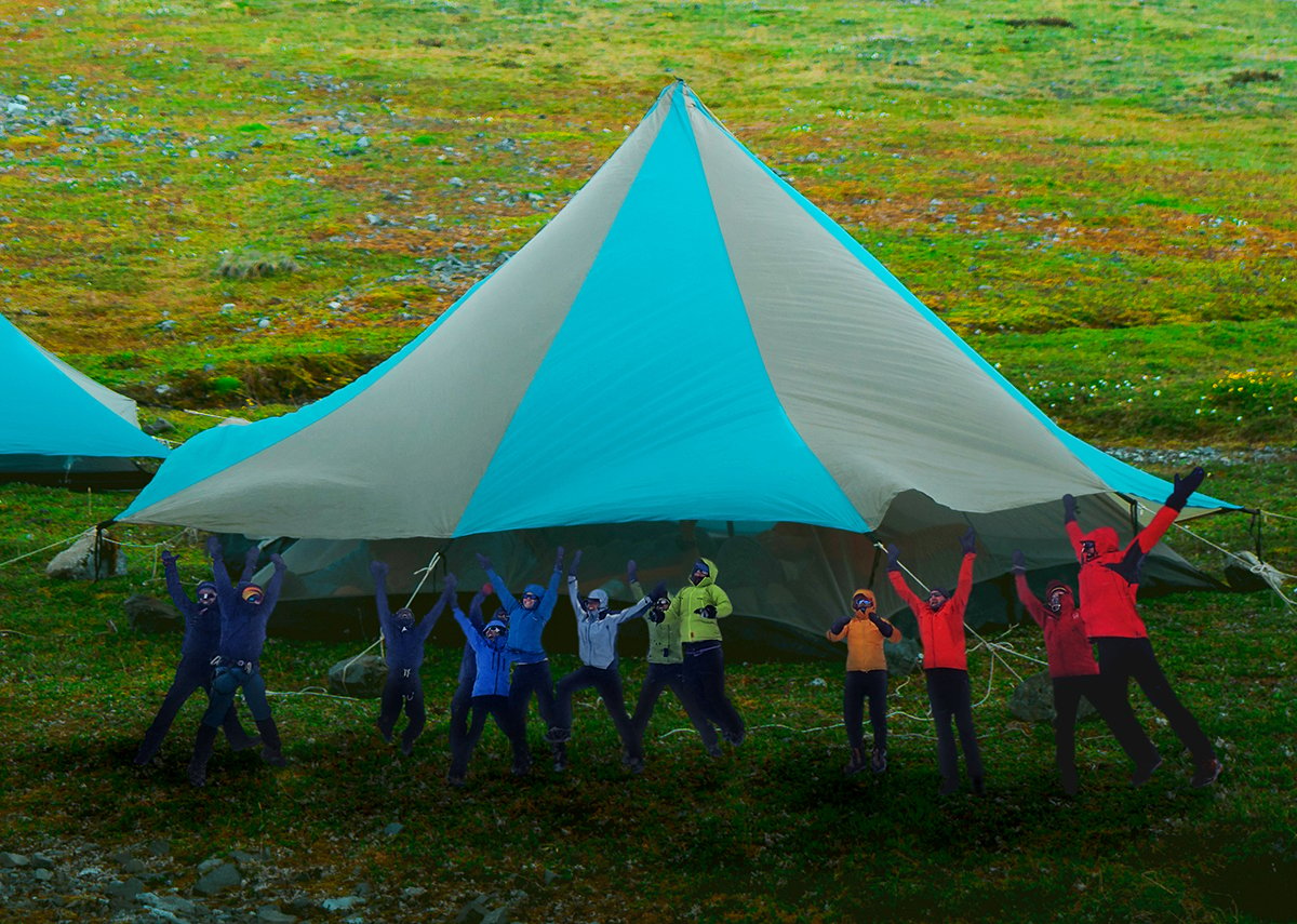 People in colorful jackets jump into the air with raised arms in front of a giant blue and tan tent