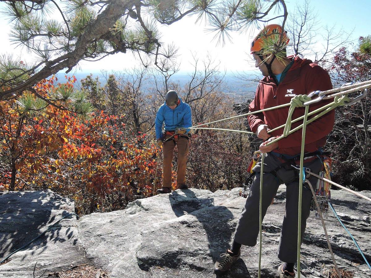 rock climbers connected by a harness at the top of a climb with fall leaves behind and pine branch overhanging