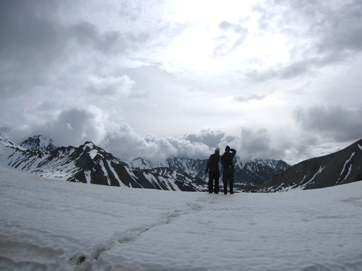 two silhouetted figures stand on a snowfield in Alaska, looking out toward mountains topped with clouds