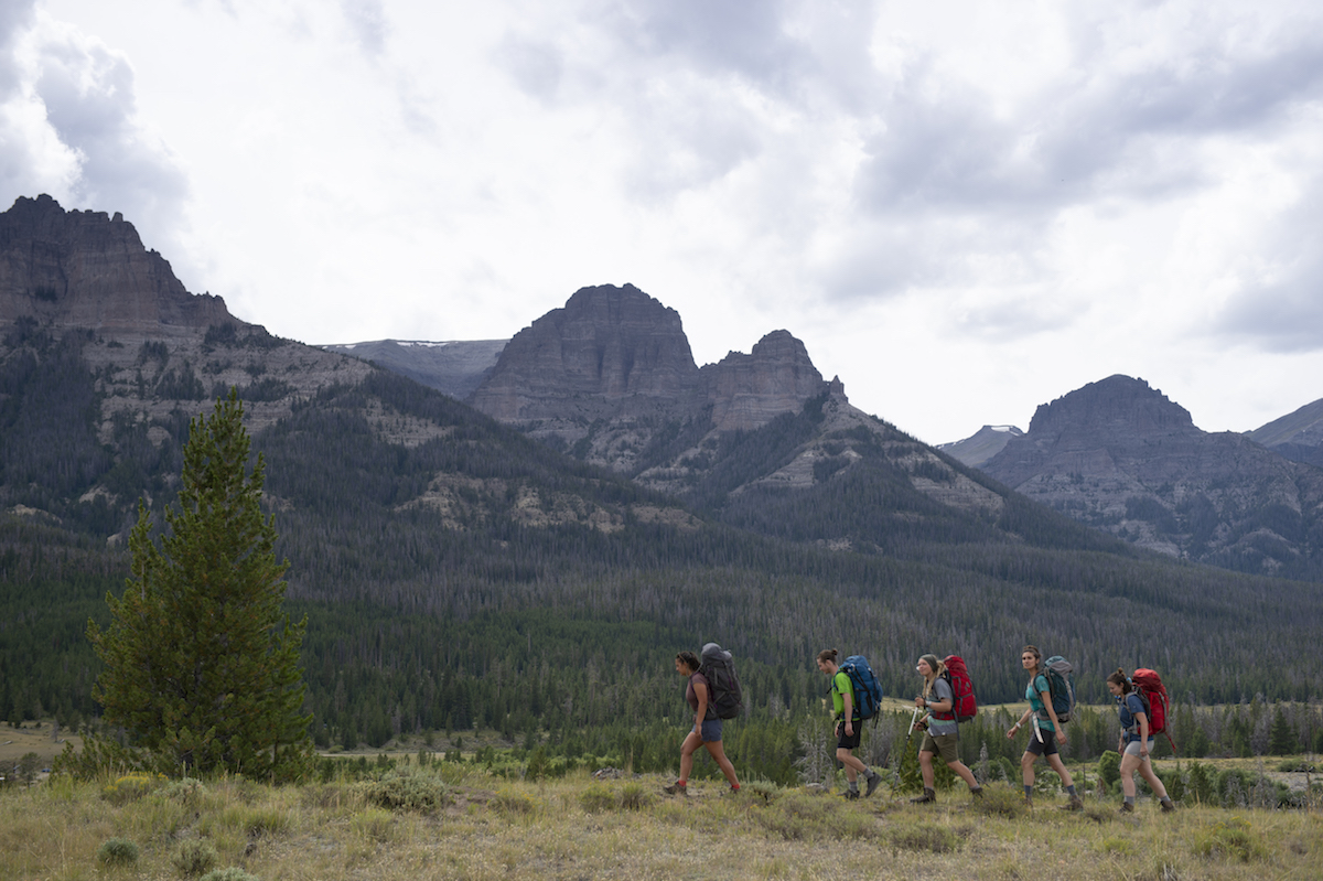 four backpackers hiking past pines and rocky peaks under cloudy sky