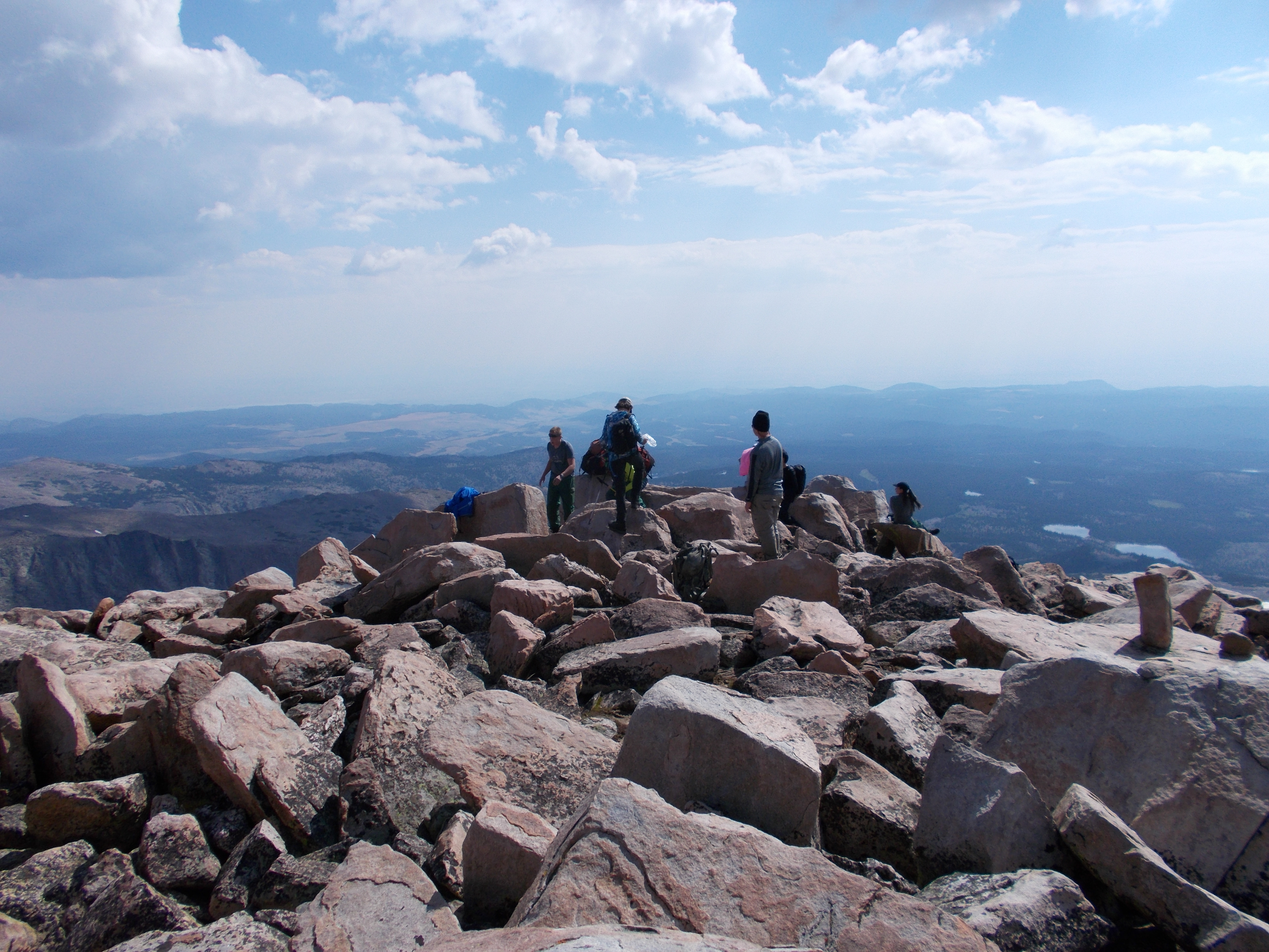 A group of four backpackers cross a boulder field with a large mountain range in front of them