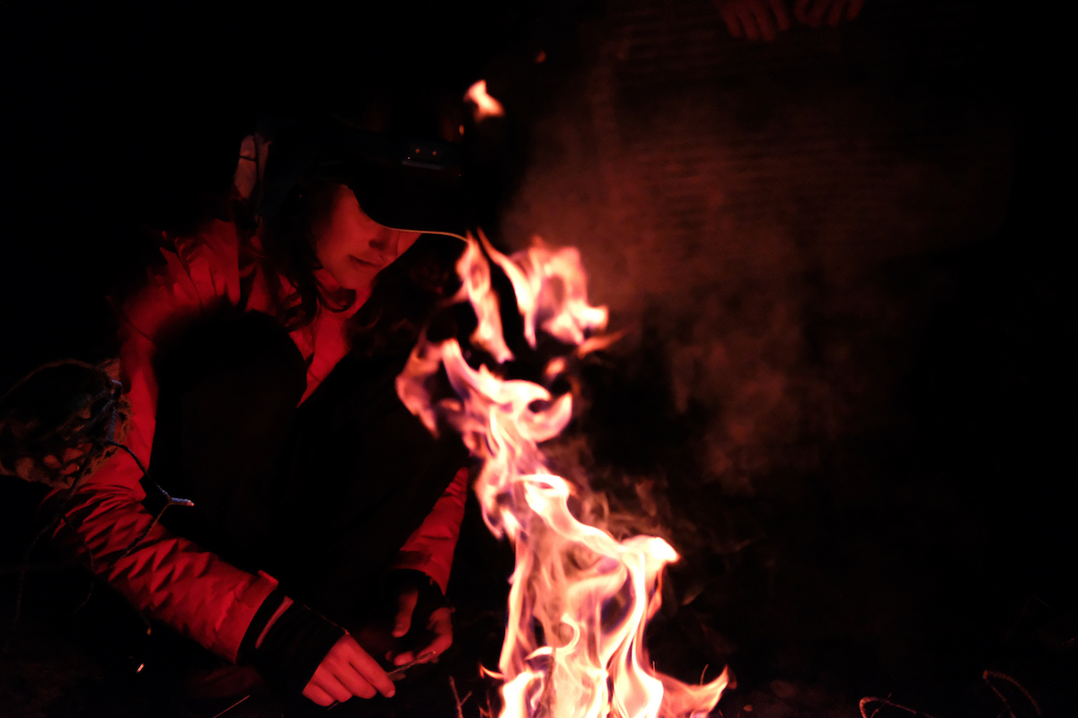 person wearing baseball cap tends campfire at night