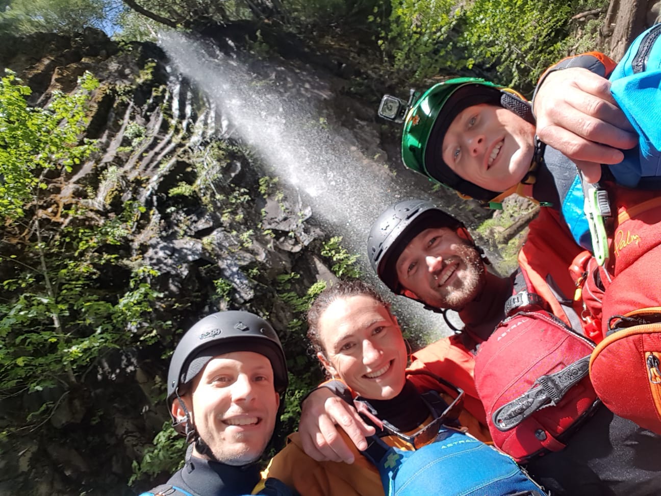 Four smiling people standing in front of a waterfall wearing lifejackets and helmets