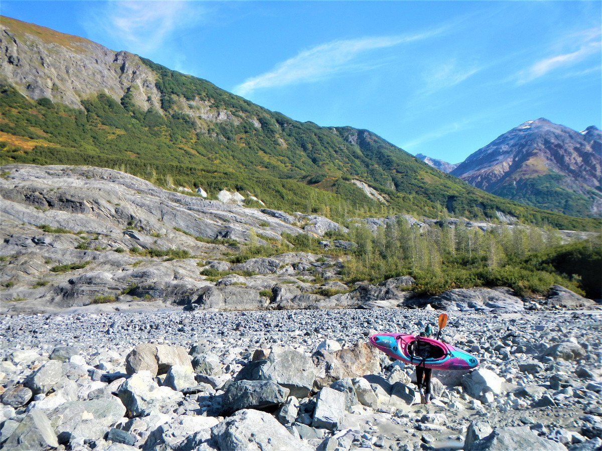 Woman carries pink and blue kayak across rocks while portaging on the Alsek River