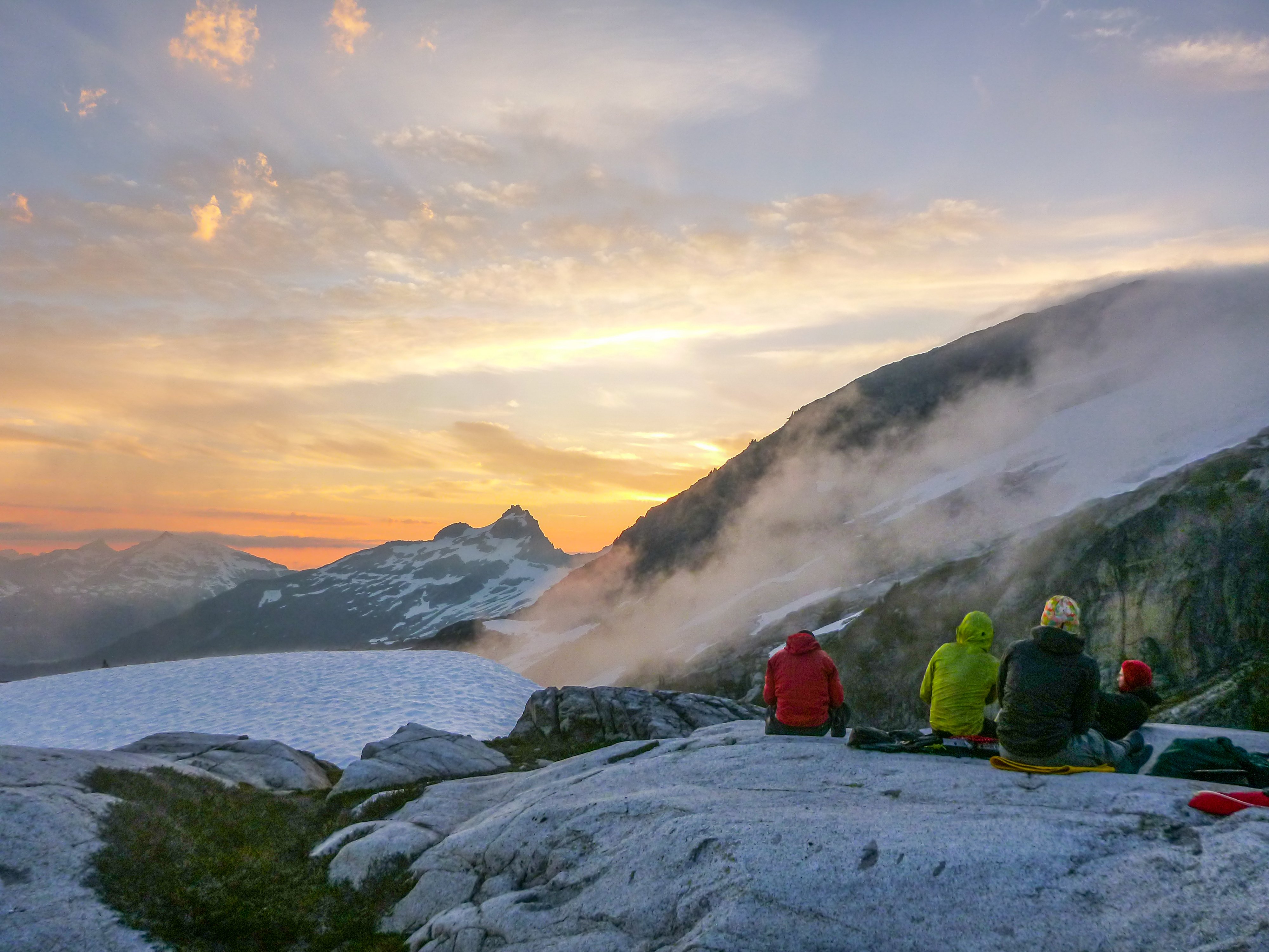 NOLS students sit on a rocky outcrop in the mountains of the Pacific Northwest at sunset with wisps of clouds