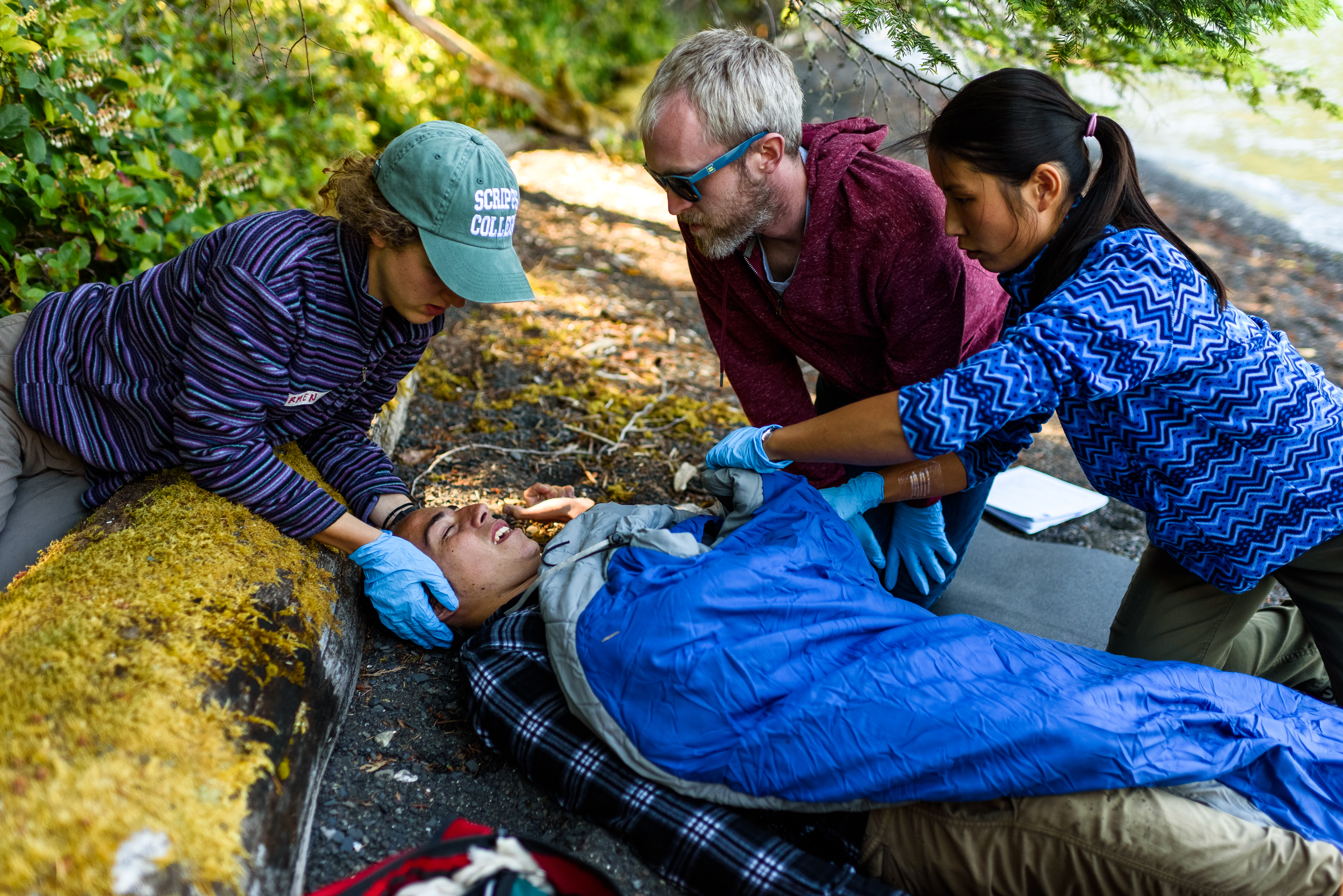 A group of three rescuers wrap a hypothermic patient into a sleeping bag to create a hypothermia wrap. They are in a forrest.