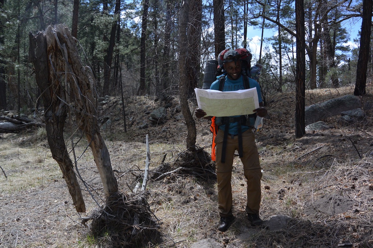 smiling NOLS student wearing backpack pauses to read a map while hiking through pines in the Southwest