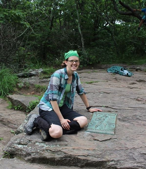 A young woman in plaid and hiking boots stops on a rock in a forest to look at a map titled Appalachian Trial.