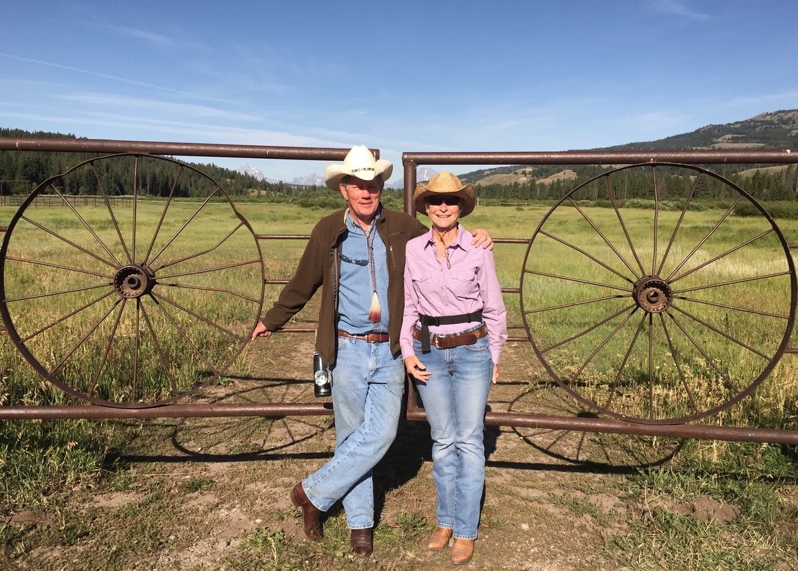 smiling couple wearing cowboy hats and boots stand in front of metal gate in Jackson, Wyoming