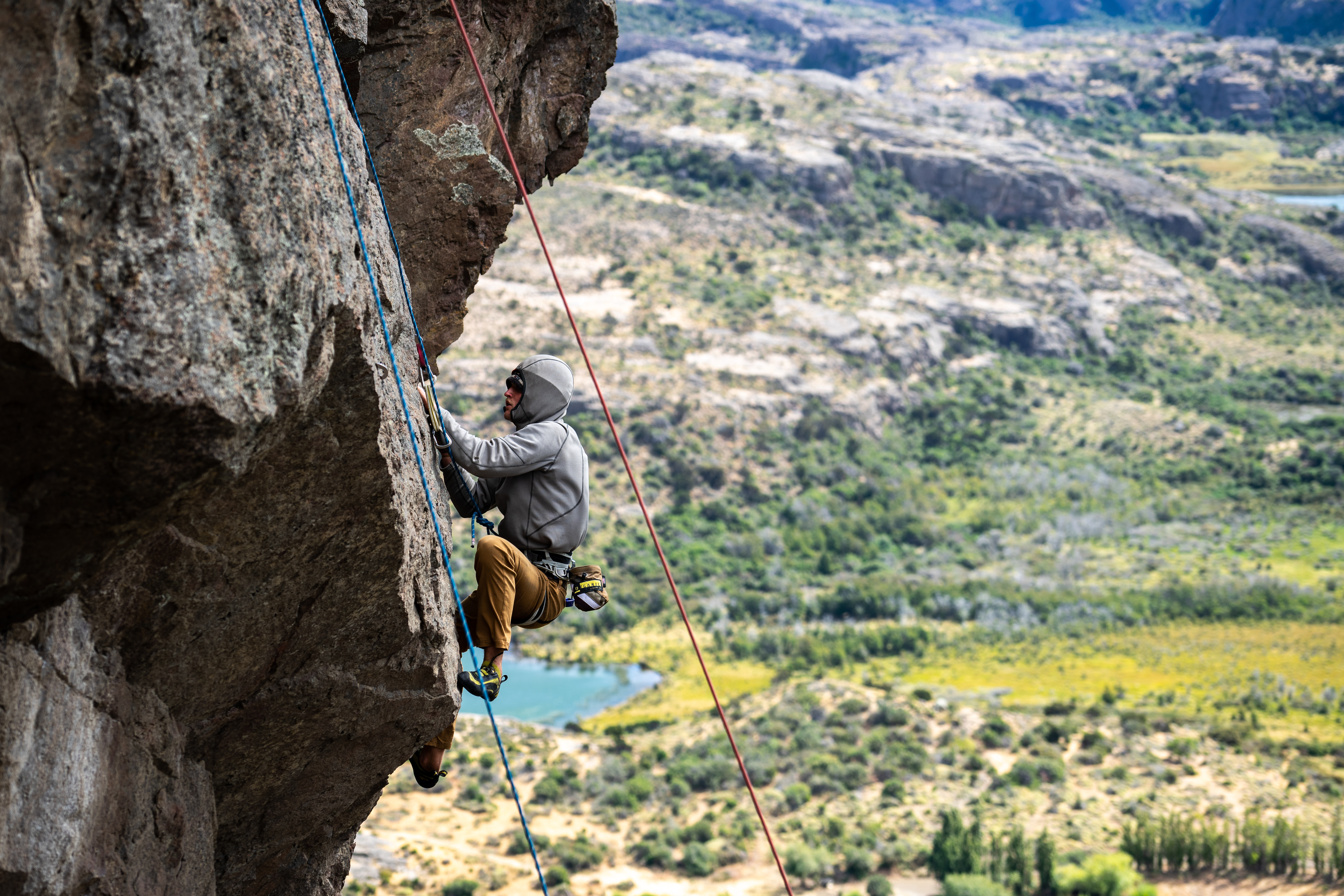 A man climbs on the ledge of a steep rock. There is a a valley of many trees and a lake below.