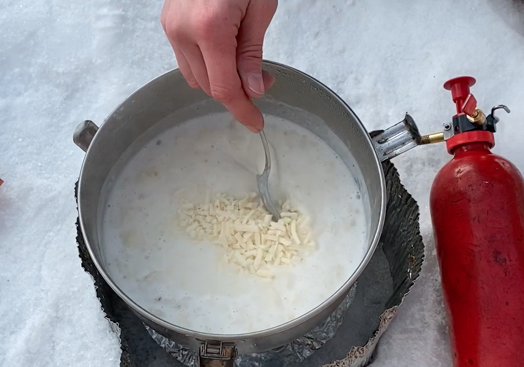 hand stirs grated cheese into potato soup over a WhisperLite stove